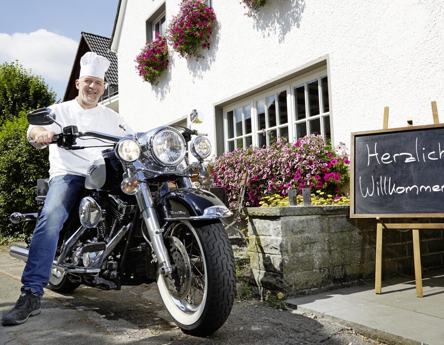 Koch vor dem Haus Dumicketal mit Motorrad, Blumen und Angebotstafel