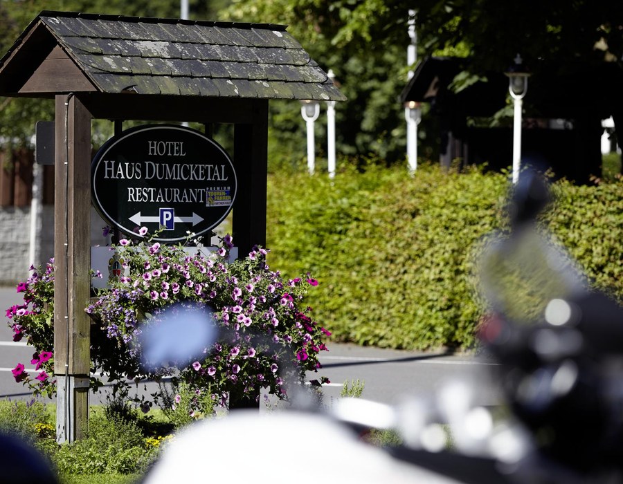 Schild des Haus Dumicketal vor dem Hotelgarten mit Blumen und Motorrad im Vordergrund